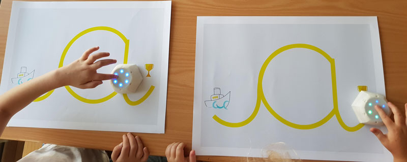 Figure 1: Robot-assisted writing activity with Cellulo: Children interact with the haptic-enabled Cellulo robots on maps displaying the grapheme of a cursive letter, where the start and end of the letter are indicated with a starting cue and a trophy respectively. After three different sub-activities where the robots provide a range of sensory information, a final team activity in the form of a guessing game is played.
